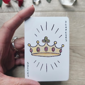 May include: A white card featuring a gold crown with red gems and radiating lines. The words "HONNEURS" and "SUCCES" are printed vertically on the card. The card is held by a person with red nail polish.