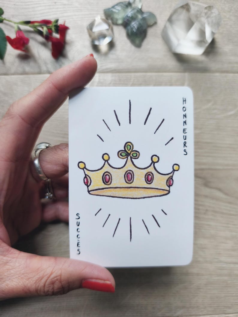 May include: A white card featuring a gold crown with red gems and radiating lines. The words "HONNEURS" and "SUCCES" are printed vertically on the card. The card is held by a person with red nail polish.