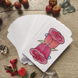 May include: A deck of white cards with a red rose illustration, fanned out on a wooden surface. The image includes red berries, a red flower, and decorative crystal objects. The rose illustration has pink and red petals with green leaves.