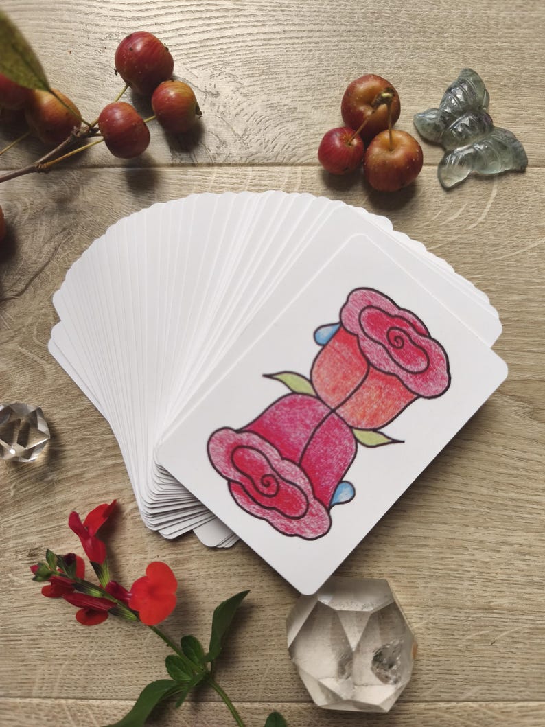 May include: A deck of white cards with a red rose illustration, fanned out on a wooden surface. The image includes red berries, a red flower, and decorative crystal objects. The rose illustration has pink and red petals with green leaves.