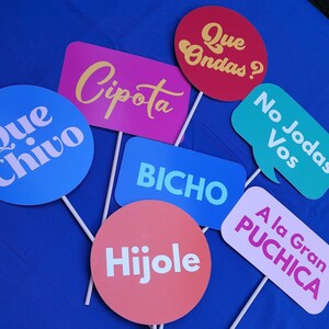 May include: A set of colorful cardboard signs with Spanish phrases, including "Que Ondas?", "Cipota", "Que Chivo", "No Jodas Vos", "Bicho", "Hijole", and "A la Gran Puchica".