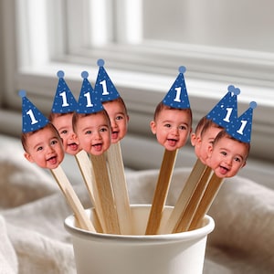 May include: A set of party picks featuring cutouts of a baby's face wearing a blue cone-shaped party hat with white polka dots and the number one printed on the front. Each face cutout is attached to a flat wooden stick, all held in a white cup.
