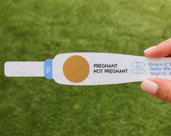 May include: A white paper pregnancy test game card featuring a circular scratch-off area, the text PREGNANT and NOT PREGNANT, and the printed text Grace & Tom's Baby Shower Sept 17, 2027, with a blue TF logo on the left side.