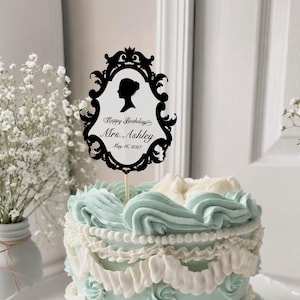 May include: A decorated cake with light blue and white frosting, topped with a black silhouette cake topper. The topper has a decorative frame with the text "Happy Birthday Mrs. Ashley May 16, 2027".