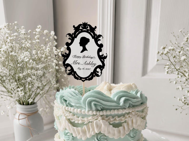 May include: A decorated cake with light blue and white frosting, topped with a black silhouette cake topper. The topper has a decorative frame with the text "Happy Birthday Mrs. Ashley May 16, 2027".