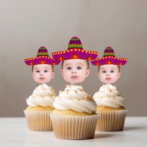 May include: Three cupcakes with white frosting and gold sprinkles, each topped with a baby's face wearing a colorful sombrero. The cupcakes are in brown paper liners, set against a neutral background.