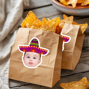 Peut inclure: Deux sacs en papier brun remplis de chips de tortilla jaune doré. Chaque sac présente un autocollant représentant le visage d'un bébé portant un sombrero coloré violet, rouge et vert avec des motifs décoratifs sur le devant de l'emballage.