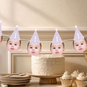 May include: A festive first birthday scene with a banner of baby faces wearing lavender party hats with the number 1. A frosted cake sits on a cake stand, surrounded by cupcakes, plates, and champagne flutes.