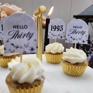 May include: Four cupcakes with white frosting and gold cupcake liners. Each cupcake has a decorative topper. One topper says "Hello Thirty", another says "1993", and two toppers say "Hello Thirty".