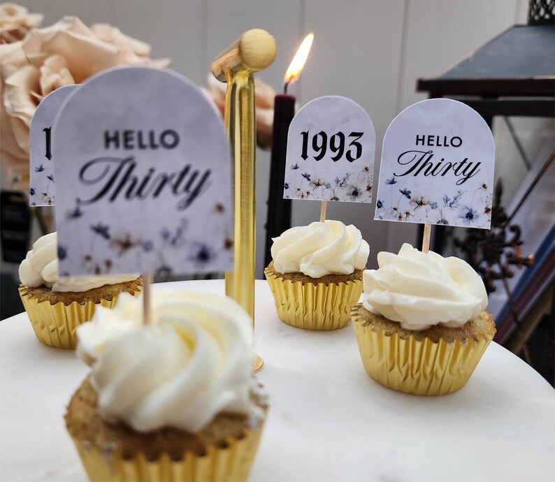 May include: Four cupcakes with white frosting and gold cupcake liners. Each cupcake has a decorative topper. One topper says "Hello Thirty", another says "1993", and two toppers say "Hello Thirty".