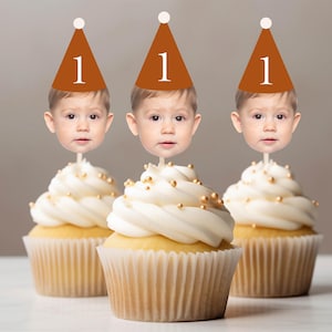 May include: Three cupcakes with white frosting and gold sprinkles. Each cupcake has a baby's face on a stick, wearing a brown party hat with the number 1.