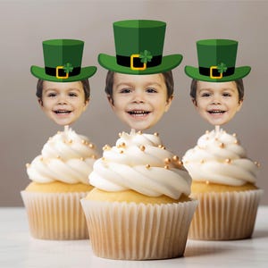 May include: Three cupcakes with white frosting and gold sprinkles. Each cupcake is topped with a photo of a child wearing a green leprechaun hat with a black band and gold buckle. The cupcakes are in paper liners.