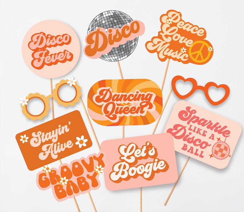 May include: A collection of retro-themed photo booth props on wooden sticks. The props feature phrases like "Disco Fever," "Dancing Queen," and "Groovy Baby" in orange and pink hues, along with a disco ball and heart-shaped glasses.