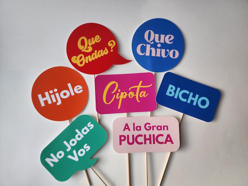 May include: A set of seven colorful photo booth props with Spanish phrases, including "Que Ondas?", "Hijole", "No Jodas Vos", "Que Chivo", "Cipota", "Bicho", and "A la Gran Puchica".