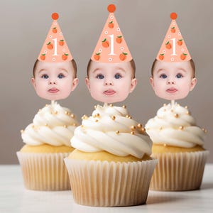Könnte beinhalten: Drei Cupcakes mit weißem Zuckerguss und goldenen Streuseln. Jeder Cupcake ist mit einem Babygesicht und einer Partyhütchen mit der Zahl "1" und Pfirsichillustrationen verziert, ideal für den ersten Geburtstag.