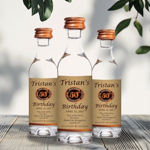 May include: Three clear glass bottles with copper-colored caps and labels that read "Tristan's 50th Birthday." The labels are tan with black and orange text. The bottles are on a wooden surface, with green leaves in the background.