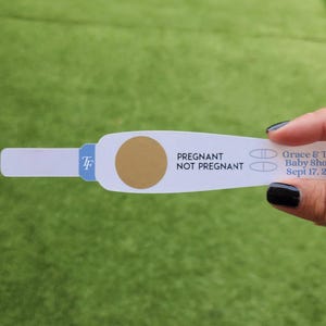 May include: A white pregnancy test with the words "Pregnant" and "Not Pregnant" printed on it. The test has a gold circle and two lines. The text "Grace & Tom's Baby Shower Sept 17, 2026" is also printed on the test.