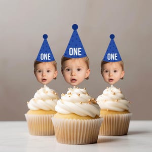 May include: Three cupcakes with white frosting and gold sprinkles, each topped with a cutout of a child's face wearing a blue party hat with white polka dots and the word ONE printed in white on the front of each hat.