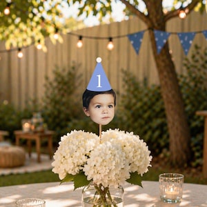 May include: A custom photo cake topper featuring a cutout of a child's face wearing a light blue party hat with the number 1 printed in white, attached to a wooden stick and displayed in a glass vase filled with white hydrangea flowers.