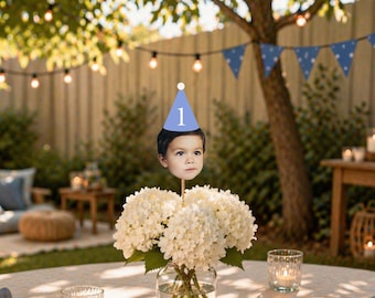 May include: A custom photo cake topper featuring a cutout of a child's face wearing a light blue party hat with the number 1 printed in white, attached to a wooden stick and displayed in a glass vase filled with white hydrangea flowers.