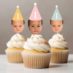 May include: Three cupcakes with white frosting and gold sprinkles topped with a cupcake topper featuring a child's face wearing a party hat with the number "1" on it.