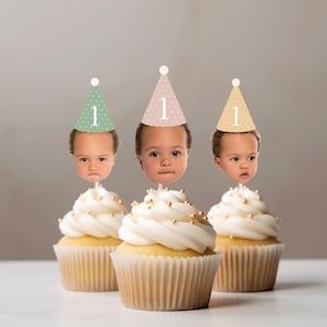 May include: Three cupcakes with white frosting and gold sprinkles, each topped with a cutout of a baby's face wearing a conical party hat. The hats are sage green, dusty pink, and beige, each featuring a white polka dot pattern and the number 1.
