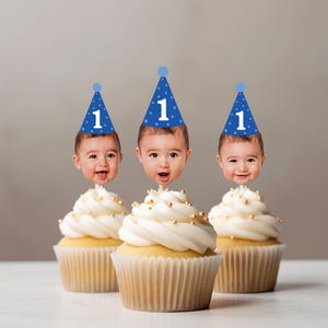 May include: Three cupcakes with white frosting and gold sprinkles, each topped with a cutout of a baby's face wearing a blue party hat with a white number 1 printed on it. The cupcakes are in light-colored paper liners and arranged in a row.