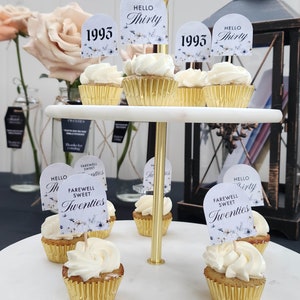 May include: A two-tiered cake stand with cupcakes decorated with white frosting and floral toppers. The toppers have text that says "Hello Thirty", "1993", and "Farewell Sweet Twenties".