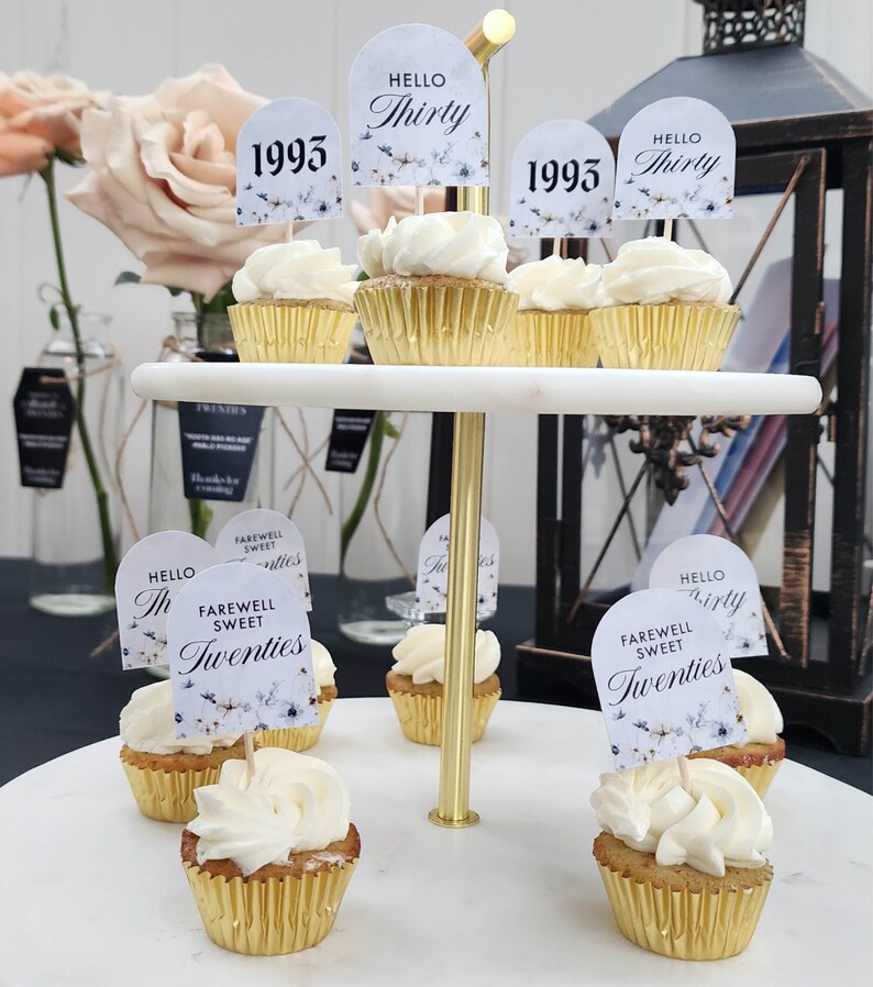 May include: A two-tiered cake stand with cupcakes decorated with white frosting and floral toppers. The toppers have text that says "Hello Thirty", "1993", and "Farewell Sweet Twenties".