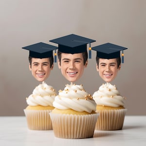 Può includere: Tre cupcake con glassa bianca, spruzzi dorati e un volto sorridente con cappello di laurea. I cupcake sono in pirottini di carta, su sfondo neutro.