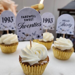 May include: A close-up of cupcakes decorated with white frosting and topped with small flags. The flags have different messages, including "Farewell Sweet Twenties", "1995", and "Hello Thirty".