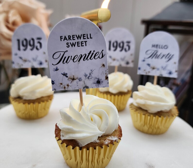 May include: A close-up of cupcakes decorated with white frosting and topped with small flags. The flags have different messages, including "Farewell Sweet Twenties", "1995", and "Hello Thirty".
