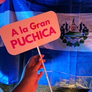 May include: A pink sign with the text "A la Gran PUCHICA" in front of a blue and white flag with the coat of arms of El Salvador.