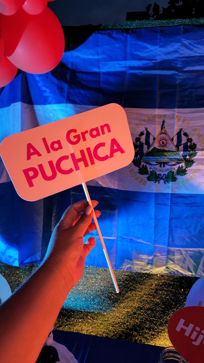 May include: A pink sign with the text "A la Gran PUCHICA" in front of a blue and white flag with the coat of arms of El Salvador.