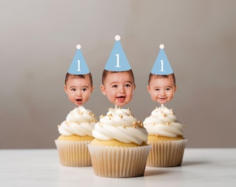 May include: Three cupcakes with white frosting, gold sprinkles, and baby face toppers wearing light blue party hats with the number 1. The cupcakes sit on a white surface against a neutral background, perfect for a first birthday celebration.