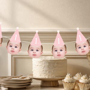 May include: A decorative party banner featuring a repeating sequence of baby faces wearing pink cone-shaped party hats. Each hat is adorned with a white number one and a small white pom-pom at the top. The faces are strung together on a thin white cord.
