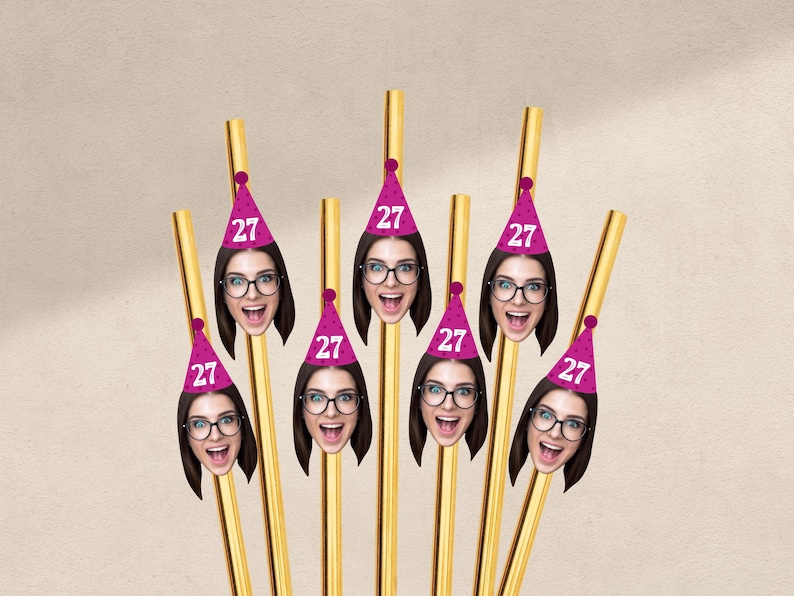May include: Gold-colored straws with attached party hats featuring a woman's face and the number "27". The hats are pink with a white number. The woman is wearing glasses and has a surprised expression.