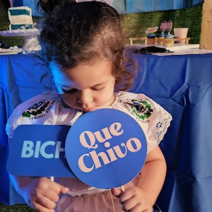 May include: A young girl in a white and blue dress holds two blue signs. One sign says "Que Chivo" and the other says "BICH". The girl is standing in front of a blue tablecloth and a flag of El Salvador.