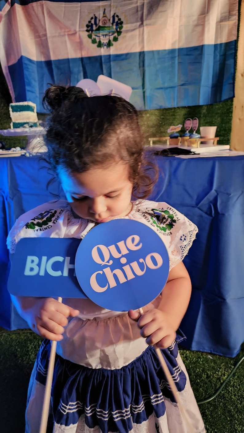 May include: A young girl in a white and blue dress holds two blue signs. One sign says "Que Chivo" and the other says "BICH". The girl is standing in front of a blue tablecloth and a flag of El Salvador.