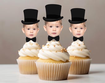 May include: Three cupcakes with light-coloured cake bases and white frosting topped with small gold sprinkles. Each cupcake features a cutout of a baby face wearing a black top hat and a black bow tie, creating a whimsical decorative topper set.