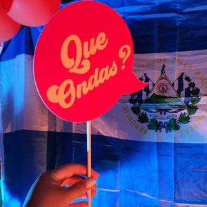 May include: A red and yellow speech bubble with the text "Que Ondas?" in front of a blue and white Nicaraguan flag.