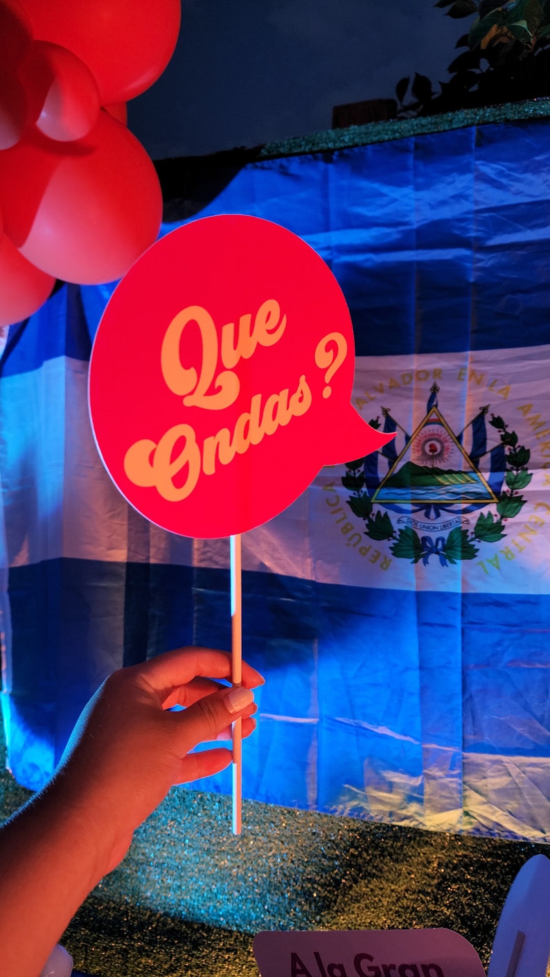 May include: A red and yellow speech bubble with the text "Que Ondas?" in front of a blue and white Nicaraguan flag.