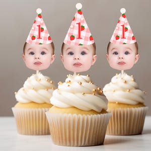 May include: Three cupcakes with white frosting and gold sprinkles. Each cupcake has a baby face topper wearing a pink gingham party hat with a strawberry and the number 1.