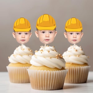 May include: Three cupcakes with white frosting and gold sprinkles, each topped with a paper cutout of a toddler face wearing a yellow construction hard hat. The cupcakes are in light-colored paper liners and arranged in a row on a flat surface.