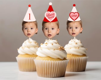 May include: Three cupcakes topped with white frosting and gold sprinkles, each featuring a cutout of a baby's face wearing a party hat. The hats display the text 1, sweet heart, and one, with red and pink heart accents on a light background.