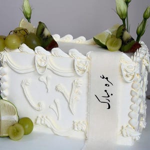 May include: A rectangular white cake decorated with white frosting, green grapes, kiwi slices, and lime wedges. The cake is adorned with white floral accents and a white ribbon with black text.