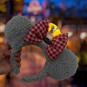 May include: A gray fuzzy headband with two round ears and a large red and white gingham bow. A yellow light-up chef hat sits in the center of the bow, featuring a silhouette of a rat holding a spoon and a tag that reads Remy's RATATOUILLE ADVENTURE.