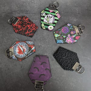 May include: Six different coffin-shaped keychains with various designs. The keychains are black, green, purple, and red with different graphic designs. The keychains are on a grey background.