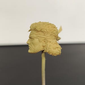 Peut inclure: Une t&ecirc;te de figurine en r&eacute;sine beige non peinte repr&eacute;sentant un personnage aux cheveux courts textur&eacute;s, portant un bandeau et une barbe courte. La t&ecirc;te est mont&eacute;e sur une fine tige verticale, offrant un profil d&eacute;taill&eacute; du visage et des cheveux.