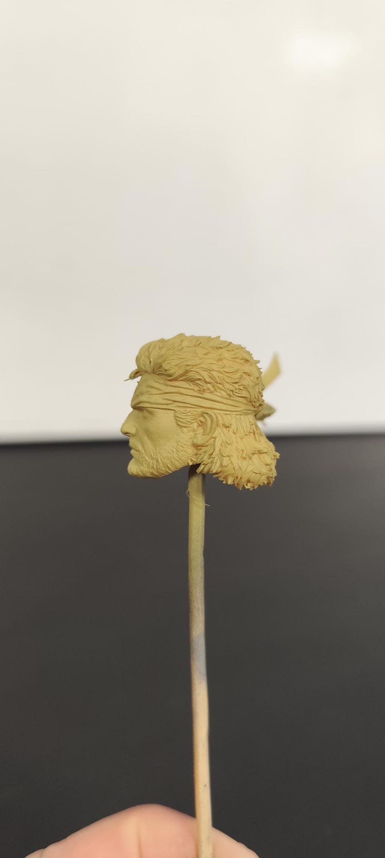 Peut inclure: Une t&ecirc;te de figurine en r&eacute;sine beige non peinte repr&eacute;sentant un personnage aux cheveux courts textur&eacute;s, portant un bandeau et une barbe courte. La t&ecirc;te est mont&eacute;e sur une fine tige verticale, offrant un profil d&eacute;taill&eacute; du visage et des cheveux.